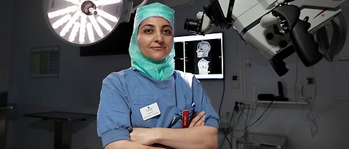 Elham Rostami står med armarna i kors, iklädd operationskläder i en operationssal. En skärm med hjärnavbildningar syns i bakgrunden. 