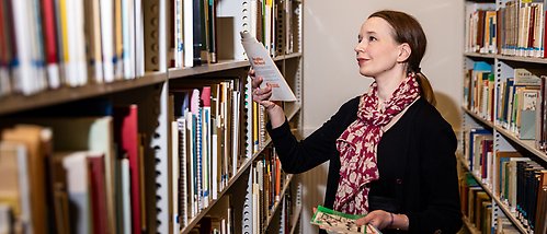 Sara Backman Prytz står mellan två hyllor i ett arkiv och plockar ut tidningar från Hem och skola. 