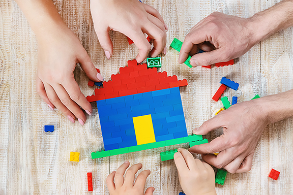 Tre par händer bygger tillsammans ett färgglatt hus i lego. 