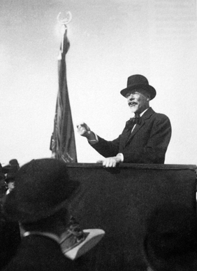 Svartvit bild av person i kostym och hatt som håller tal, i bakgrunden hänger en fana.