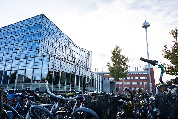 Vid cykelparkeringen utanför campus Ångström. En stor hög glasfasad i bakgrunden med texten Campus Ånströmlaboratoriet.