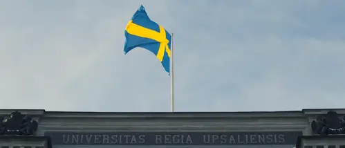 Svenska flaggan i topp ovanpå universitetshusets tak med texten "Universitas regia upsaliensis" på fasaden