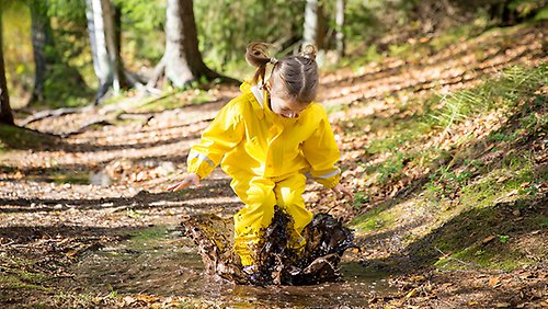 A child in yellow rain clothes jumping in a puddle of mud.