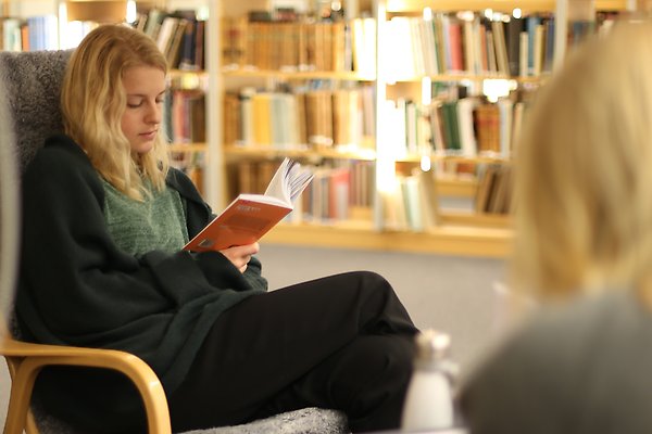 Läsande kvinna i fåtölj framför bokhylla.