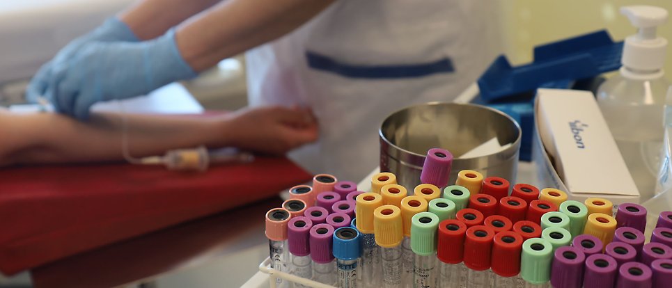 Test tubes with lids in different colors in the foreground and a hand taking a blood sample on an arm in the background.