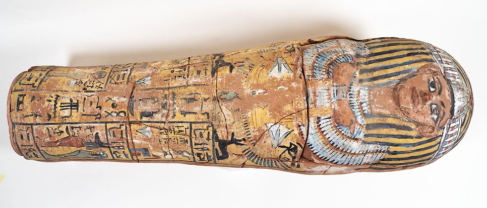 The entire sarcophagus, lying on a white table. It is made of clay and painted in different colors, a clear face with big eyes at the top.