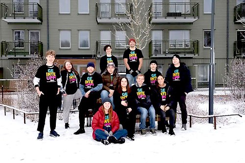 Gruppbild på unga människor som står framför ett flerfamiljshus. De har alla likadana svarta t-shirts på sig med texten "Ge inte upp". 