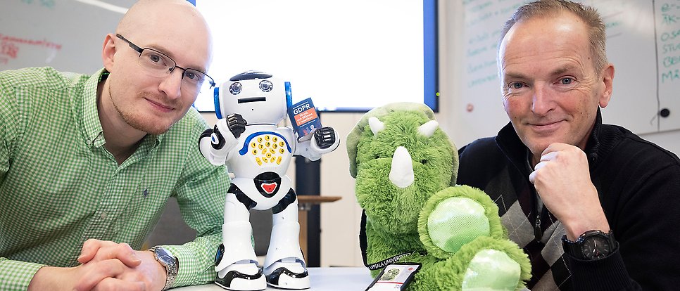 Group photo with two people, a robot toy and a green stuffed animal.