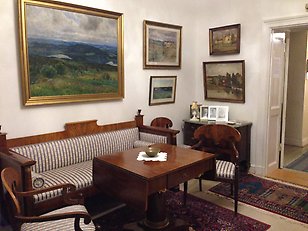 Ett rum med gammaldags inredning. Soffa och stolar i mörkt trä med en stor oljemålning med guldram ovanför soffan. Flertalet mindre målningar på väggarna. 