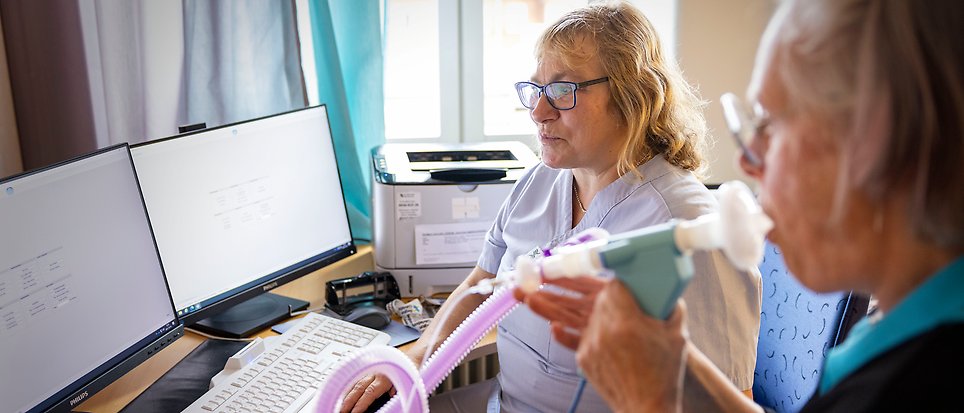 En äldre kvinnlig patient blåser i ett mätinstrument medan en sköterska tittar på.