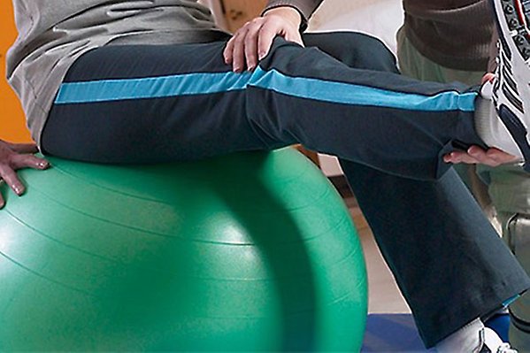 En rehabiliteringspatient sitter på en boll och fysioterapeuten hjälper med att sträcka ut benet.