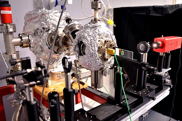 Experimental set-up for optical measurements. A light source is switched on and shines through an aperture onto a sample. Two stainless steel chambers packed in aluminium foil as well as several pipes and cables can be seen behind the optical line.
