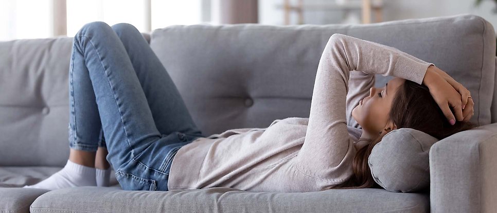 Kvinna iklädd jeans och ljus kofta ligger i grå soffa.