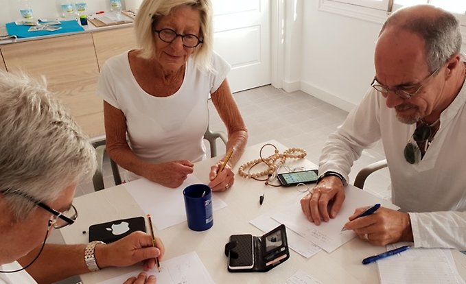 En grupp äldre personer sitter vid ett bord med papper och pennor samt telefoner framför sig.