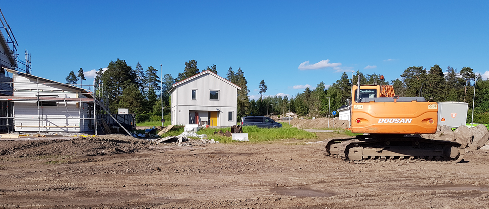 En grävmaskin står i förgrunden med bar jord runt omkring. I bakgrunden står ett nybyggt hus och till vänster i bild skymtar en byggställning. 