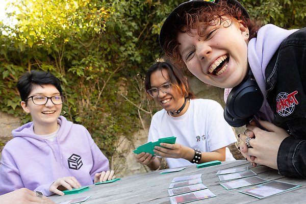 Three students sitting outdoors at a table playing cards.